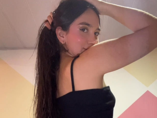 HannaCarlton with long brown hair, black strap top, holding hair up, looking over shoulder, pink blush on cheeks, right arm raised, subtle smile, indoor ceiling visible.
