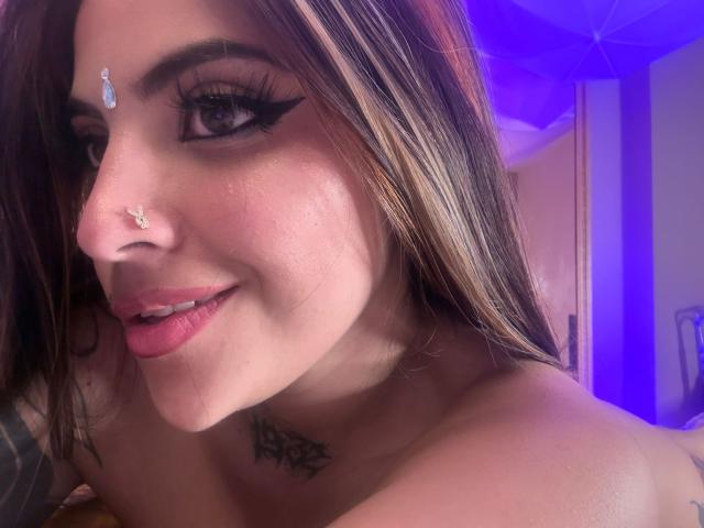 CoriPaige met bruin haar, een neusring en zwarte eyeliner, lachend met roze lippen, met een tatoeage op haar borst en een diamantversiering op haar voorhoofd..