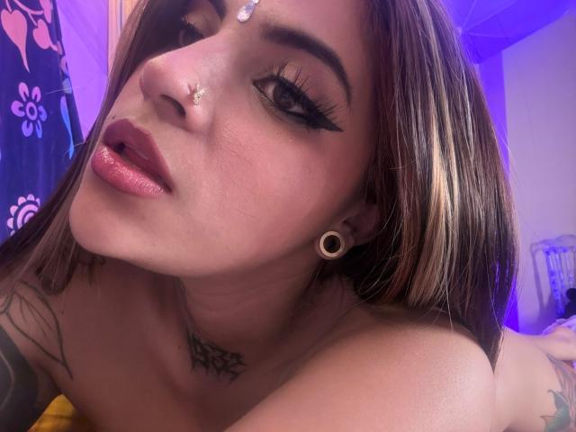 CoriPaige has glossy pink lips, dark eyeliner, and a nose ring. She's topless with tattoos, black ear gauges, and a diamond forehead jewel. Her brown hair is highlighted.