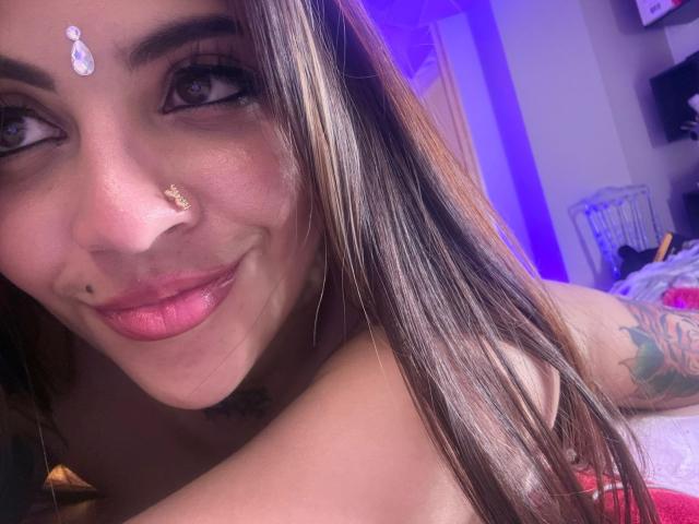 CoriPaige's face, smiling with pink lips, nose adorned with a gold septum ring and white teardrop, brown hair, tattoo on upper arm, purple lighting in background.