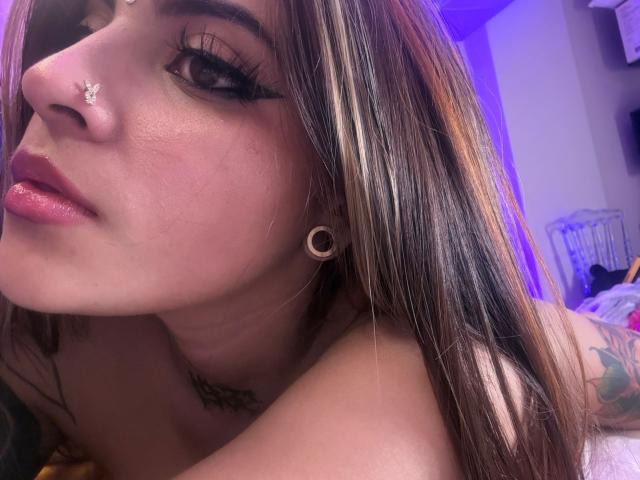CoriPaige's face with long brown hair, nose piercings, black eyeliner, and small ear gauges, showing pink lips and a tattoo on her neck. She's topless with a soft light.