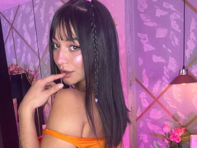 LindaWatsong with long black hair in pigtails, orange off-shoulder top, pinkish-purple lighting, finger in mouth, looking back, showing her shiny face and bare shoulder.