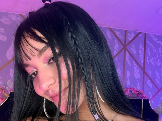 LindaWatsong with black braided hair, pink eye makeup, hoop earrings, nose ring, pouty lips, purple light, and a slight smile, showing her ass in the reflection.
