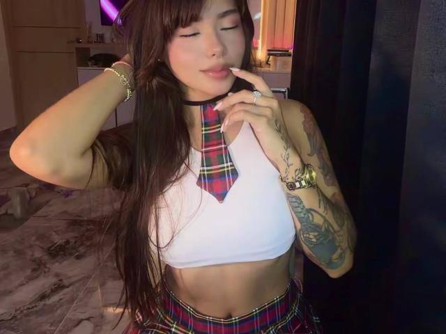 PrincessMiley with long brown hair, plaid necktie, and white crop top, showing her tattooed arm, finger to her lips, and a gold watch, wearing a matching plaid skirt.