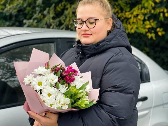 RadaRori with glasses, blonde hair in a ponytail, wearing a black puffy jacket, holding a pink-wrapped bouquet of white and pink flowers, standing next to a silver car.