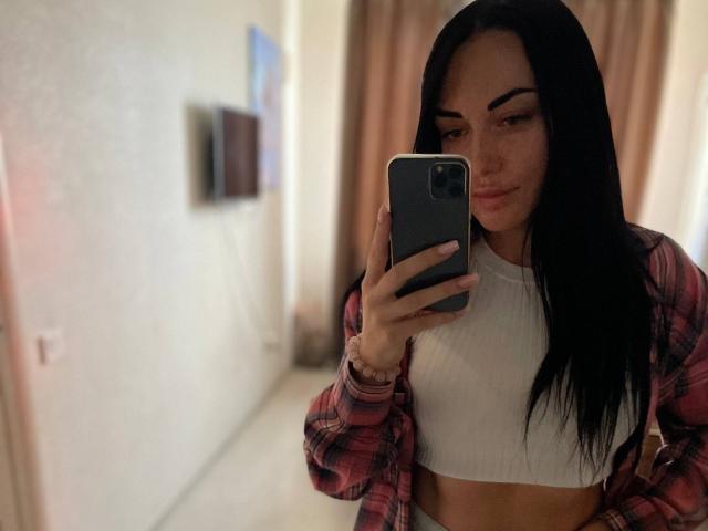 Xioni with long black hair, wearing a red plaid shirt over a white crop top, taking a mirror selfie with a confident smile, holding a grey phone.