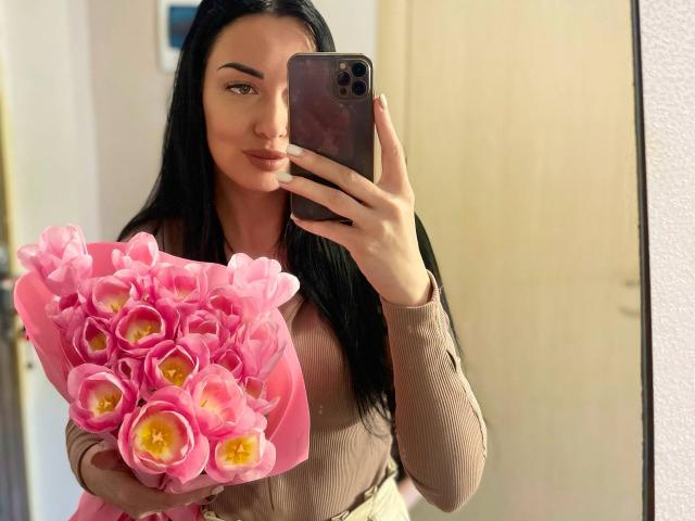 Xioni with long black hair, holding pink tulips in her right hand, taking a mirror selfie with her left. She's wearing a brown long-sleeve top, showing her tits slightly.