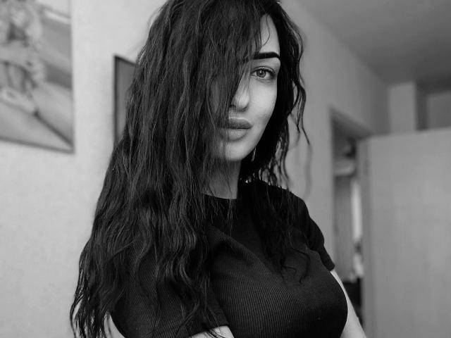 Black and white photo of Xioni with long, wavy dark hair covering one eye, wearing a black ribbed shirt, arms crossed, standing in a room, looking at camera.