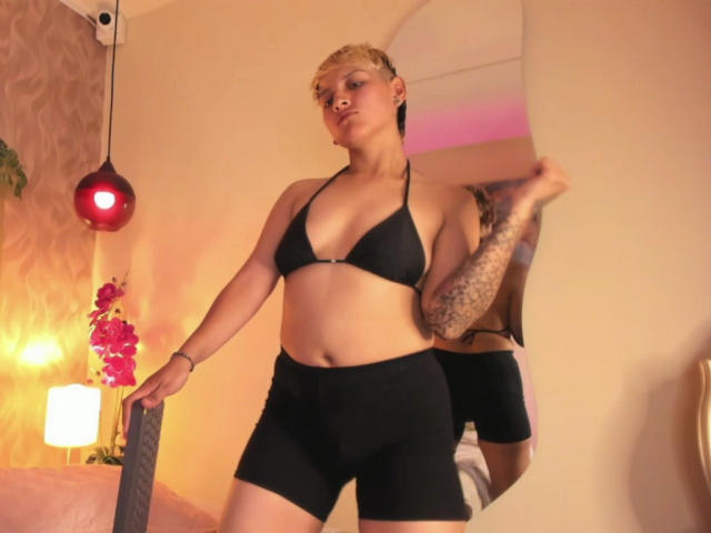 DannyRouds, short blonde hair, black bikini top, black bike shorts, tattoos on right arm, standing, holding mirror, pink orchid, red pendant light, warm yellow lighting.
