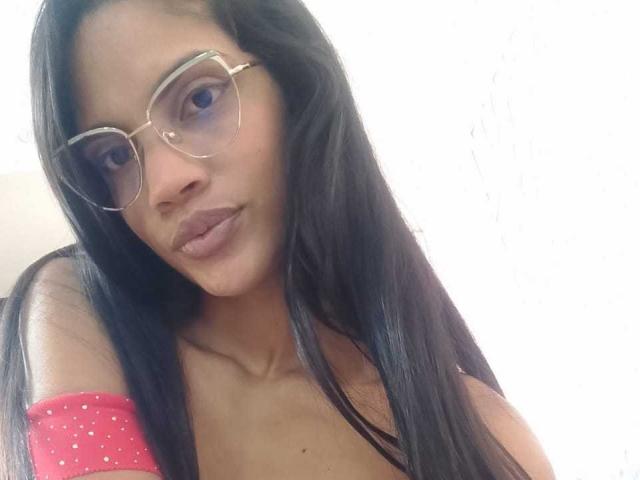 VicktoriaSunshine with long black hair, wearing glasses and a pink off-shoulder top, has full lips and a subtle smile, showing her small tits and smooth brown skin.