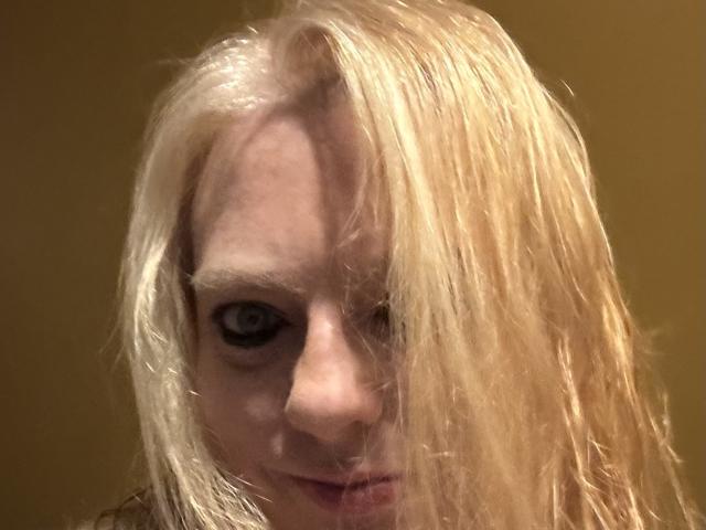CherishWilliams tiene el cabello rubio, largo y mojado que le cubre un ojo, piel pálida y ojos oscuros e intensos que miran directamente a la cámara con una sonrisa leve y misteriosa..