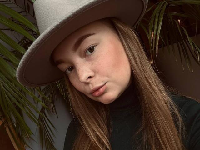 Close-up of LieMon, a young woman with fair skin and freckles, wearing a light gray floppy hat and a black turtleneck. She has long, straight brown hair, slightly parted lips, and a relaxed, slightly tilted head. Her green eyes are half-open, and she looks directly at the camera. The green palm leaves in the background are slightly blurred. The image has a natural, soft light that highlights her facial features and the texture of her hat. The composition focuses on her face and hat.