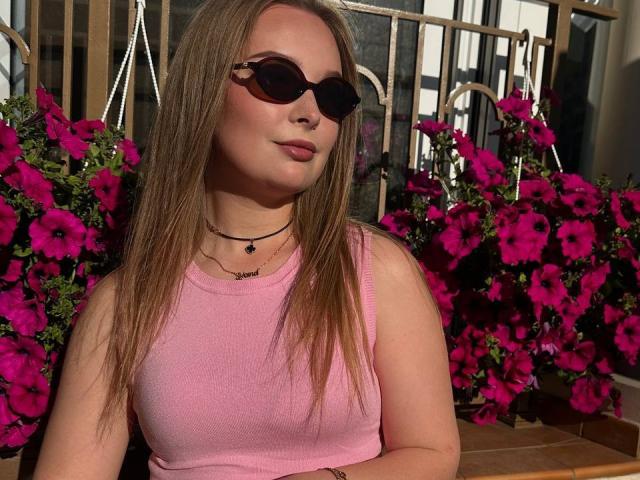 LieMon with long brown hair, wearing a pink tank top and dark sunglasses, has a subtle smile and small tits, sitting in front of bright pink flowers.