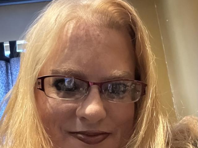 Cherish Williams tiene el cabello rubio, gafas rojas y lápiz labial oscuro, con acné visible en la frente, y está sonriendo ligeramente. Sus ojos azules reflejan la luz. Lleva una blusa marrón..