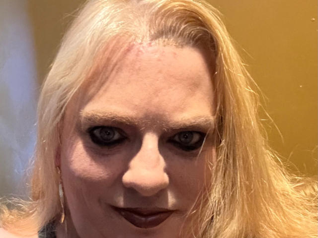 CherishWilliams has pale skin, blonde hair, dark makeup, and a slight smile. Her blue eyes are slightly squinted, and she's wearing dark lipstick. She's looking directly at the camera.