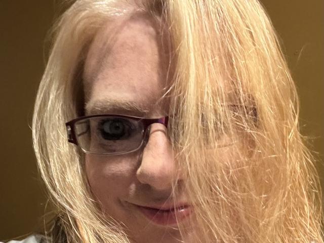Cherish Williams, met blond, rommelig haar dat een oog bedekt, draagt een donkere bril en glimlacht lichtjes, waarbij haar neus en een deel van haar lip zichtbaar zijn. Haar oog is zichtbaar en kijkt vooruit..