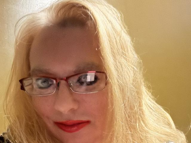 CherishWilliams has blonde hair, red glasses, and red lipstick. Her blue eyes look directly at the camera. She's wearing a black top. Her face is slightly blurry.