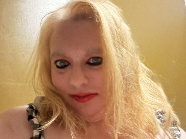 Cherish Williams, loira, com cabelo despenteado, batom vermelho e maquiagem preta nos olhos, vestindo uma blusa xadrez, olhando para a câmera com os lábios vermelhos levemente entreabertos..