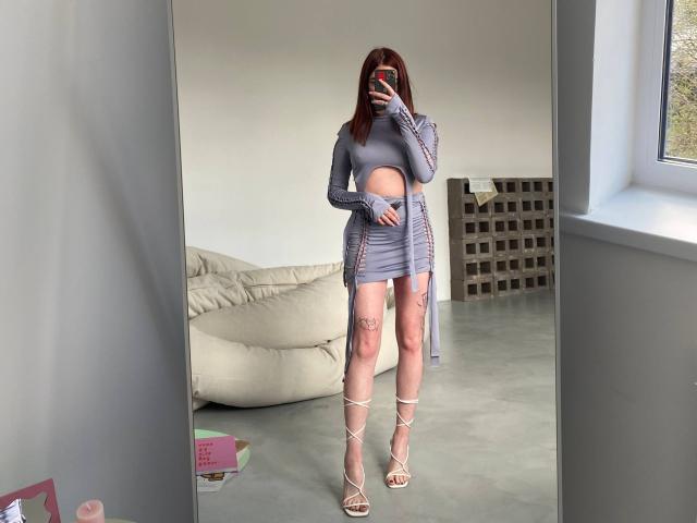 JessiBlossom in a grey cutout dress, showing her pussy through the dress, standing in front of a mirror, holding a phone, wearing strappy heels, with tattoos on her legs.