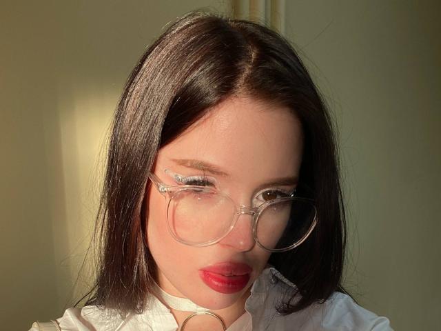 JessiBlossom has straight black hair, clear glasses, red lipstick, white shirt with collar, and a white choker. Sunlight highlights her face. Boobs not visible. Ass not visible. Pussy not visible.