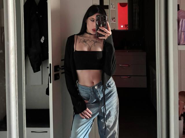 JessiBlossom with black crop top, black long sleeves, blue high-waisted jeans, tattooed chest, holding phone, mirror selfie, long black hair, red light in background.