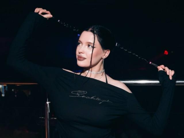 JessiBlossom with black braided pigtails, wearing an off-shoulder black top, holding her hair up, showing her tits, dark makeup, and a choker, in a dimly lit club.
