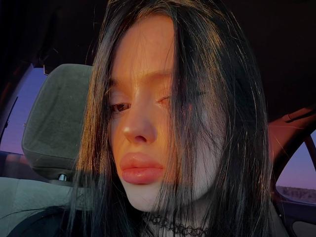 JessiBlossom's face in a car, winking with one eye, long black hair, shiny pink lips, subtle makeup, black choker, warm sunlight, relaxed expression, slight shadow on her cheek.