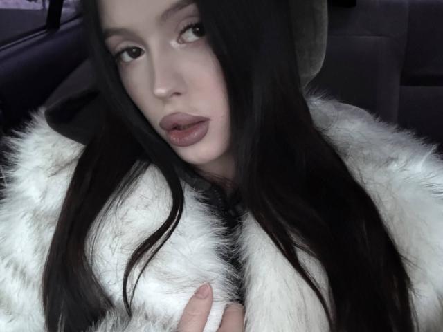 JessiBlossom with long black hair, pale skin, and full lips, wearing a white furry coat, looking at the camera with a slightly open mouth, fingers touching the coat.