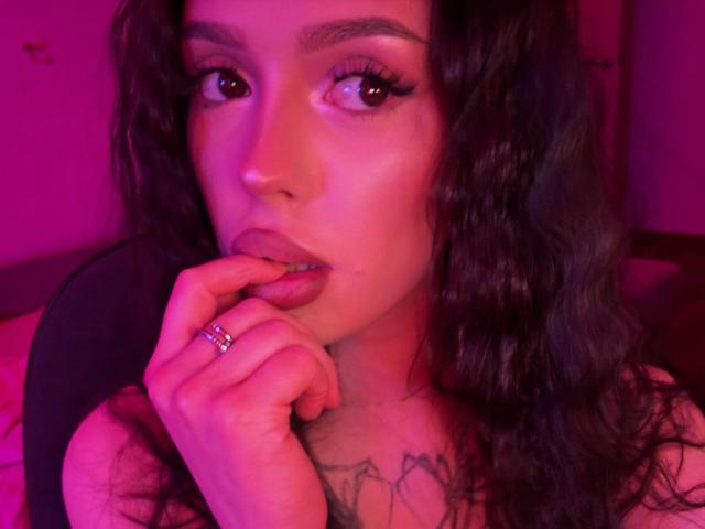 Close-up of JessiBlossom with wavy, dark brown hair, pinkish-purple light illuminating her face. She has thick, arched eyebrows, long, curled eyelashes, and slightly parted lips with her index finger touching her lower lip. She wears a silver ring on her right index finger. Her skin is smooth with a slight sheen, and there's a visible tattoo on her chest. The image has a warm, pinkish-purple hue. Her gaze is directed slightly to the right.