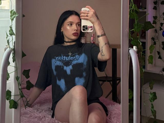 JessiBlossom with long black hair, wearing a black "Rob Zombie" t-shirt, sitting on a pink furry rug, holding a phone with tattoos and a black choker. Her ass is exposed.