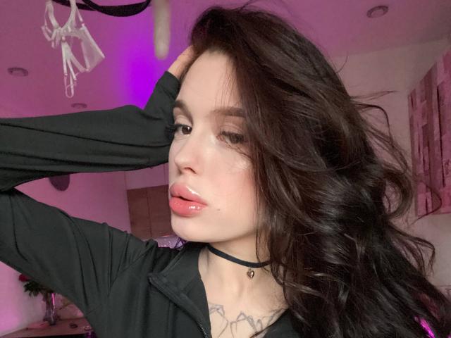 JessiBlossom with glossy pink lips, dark wavy hair, black choker, black long-sleeve top, hand in hair, tattoo on chest, pink-lit room, looking at camera.