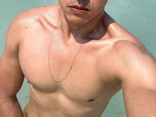 Aronpiper, shirtless, glistening with sweat, has a slim, muscular chest, small nipples, and a thin gold necklace. His face is partially visible with slightly open lips.