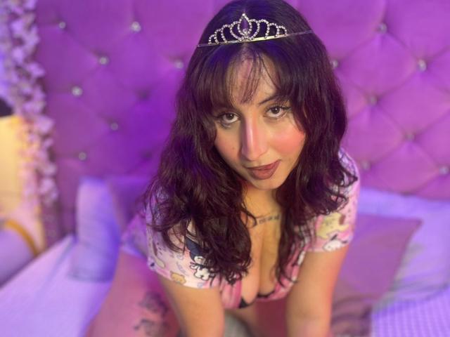 MaddieLesYoux with wavy dark hair, wearing a silver tiara, pink dress, and showing her tits, kneeling on a bed with purple and pink pillows.