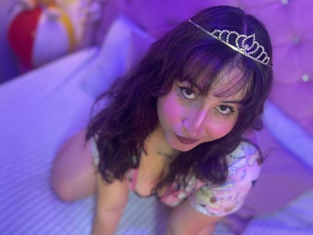 MaddieLesYoux, with wavy brown hair, wears a sparkly tiara on her head. She's on all fours on a bed with a purple-blue glow. She has dark eyebrows, large eyelashes, and is looking up at the camera with dark lipstick. She's wearing a floral-patterned top that exposes her cleavage. Her skin has a slight blush. The bed has a white, textured surface. The image has a soft, purple-blue light that highlights her face and hair.