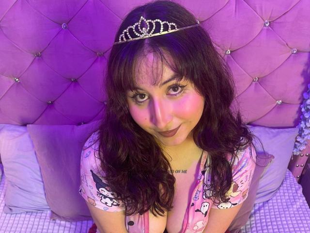 MaddieLesYoux with wavy dark hair, pink lipstick, and a sparkly tiara, wearing a pink top with cartoonish prints, sitting on a bed, looking up with a cute smile.