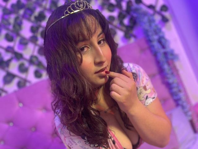 MaddieLesYoux with curly brown hair, wearing a tiara, biting her finger, showing large tits in a pink top, purple lighting, and wavy hair, looking seductively at the camera.