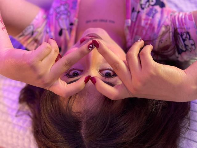 MaddieLesYoux with red nail polish, making a peace sign with fingers over her face, upside down, wearing a pink floral shirt, purple lighting, looking directly at the camera.