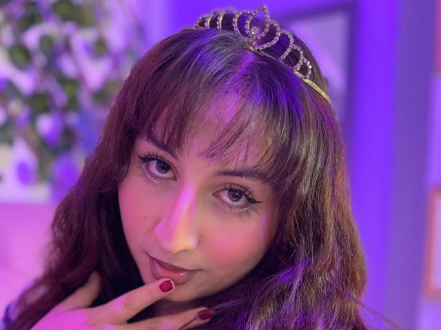 MaddieLesYoux with brown wavy hair, wearing a jeweled tiara, has purple-lit skin, red nails, and is licking her finger, looking seductive.
