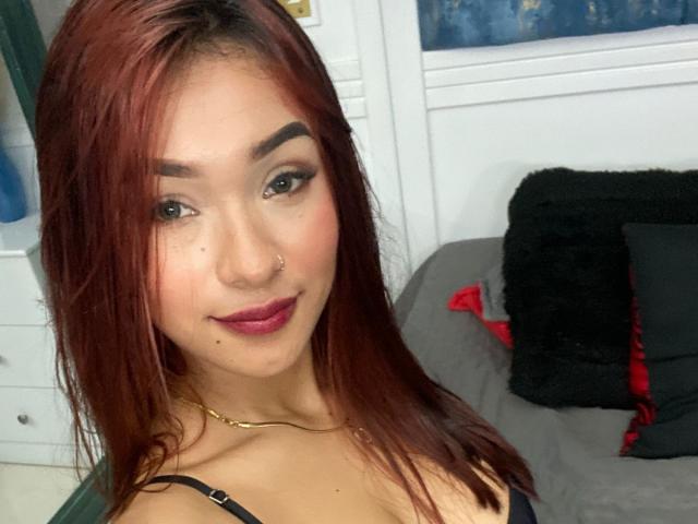 SashaHans with red hair, nose ring, and dark lipstick, smiling at the camera, showing her tits in a black strap top, wearing a gold necklace, indoors.