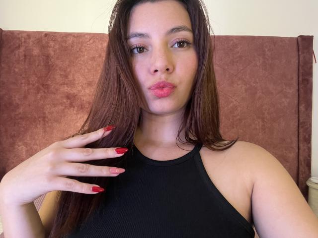 LinaFoxyHot, avec de longs cheveux bruns, des ongles rouges, un haut noir sans manches, des lèvres roses pulpeuses, une peau claire, une main sur la poitrine, une tête de lit marron, une lumière douce, regardant la caméra..
