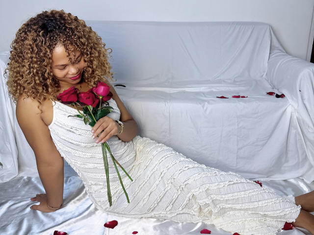 ChocolatAuLaitt with curly brown hair, white textured dress, holding red roses, sitting on white-covered couch, petals scattered, looking down with a gentle smile, soft natural light.