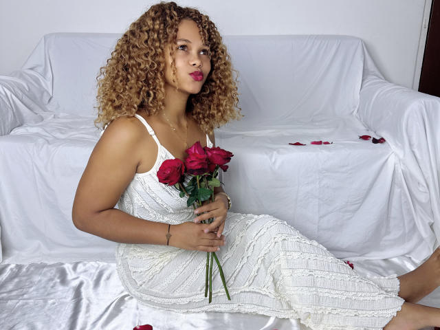ChocolatAuLaitt with curly blonde hair, wearing a white lace dress, holding red roses, sitting on white sheets, looking to the side with pink lipstick.