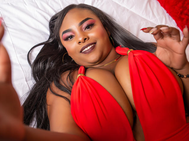 HayleyEvans with dark skin, long black hair, and pink eyeshadow, smiling at the camera. She's wearing a red halter top, highlighting her big tits. Her right hand is raised.