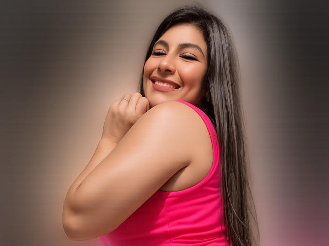 Hailin, avec de longs cheveux noirs, souriante, portant un débardeur rose, peau bronzée, tenant son menton, montrant ses fesses, et regardant par-dessus son épaule, avec une expression joyeuse..