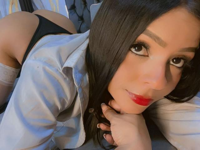 LindaFrancoo with long black hair, red lipstick, and black panties, wearing a white shirt, lying on a bed, butt up, looking at the camera, head resting on her hand.