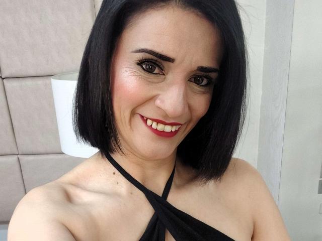 MaraRoberttss with black bob, smiling, red lipstick, black halter top, showing bare shoulders and small, visible tits. Light skin, brown eyes, and minimal makeup.