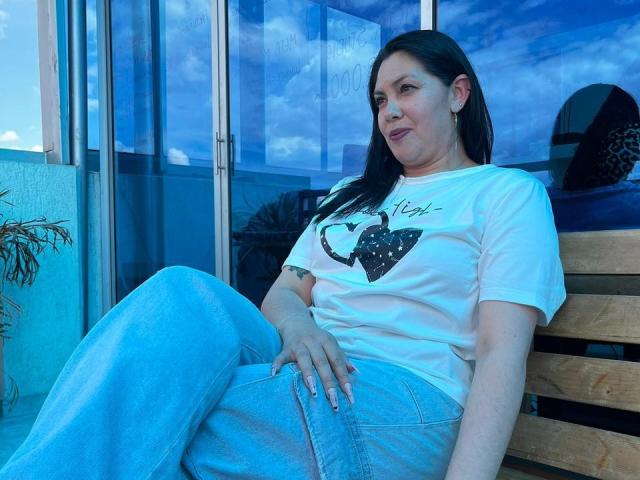 Irisbloom69 with blue jeans and white shirt, sitting on wooden bench, has black hair, light skin, small tits, and a tattoo on right hand, in blue-tinted photo.