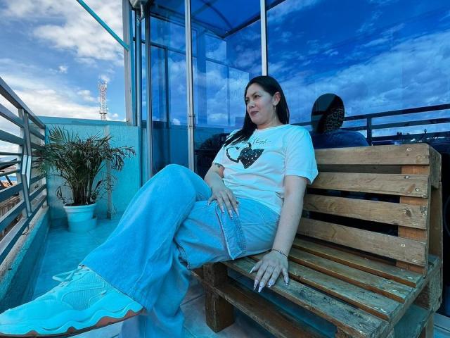 Irisbloom69 sits on a wooden bench, wearing a white tee, blue jeans, and blue sneakers, with her legs crossed, on a rooftop with glass walls and a potted plant.