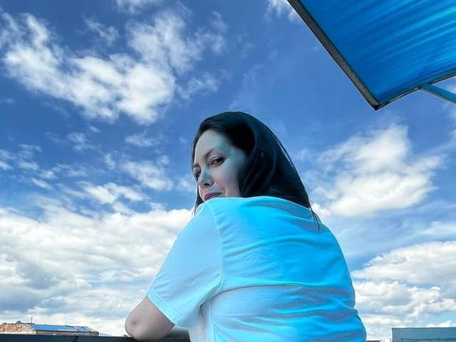 Irisbloom69 with medium tits, white shirt, black hair, looking over shoulder, under blue roof, against bright blue sky with white clouds, in daytime outdoor setting.