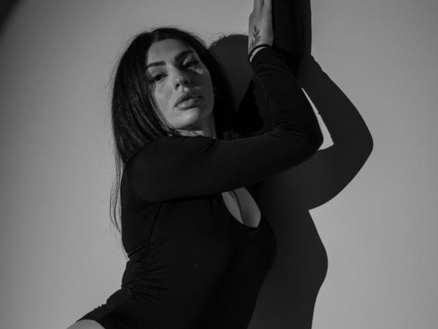 Black-and-white photo of BrainyBelle with long dark hair, wearing a tight long-sleeve top, arms raised, shadow on wall, looking seductively at camera, showing her small tits.